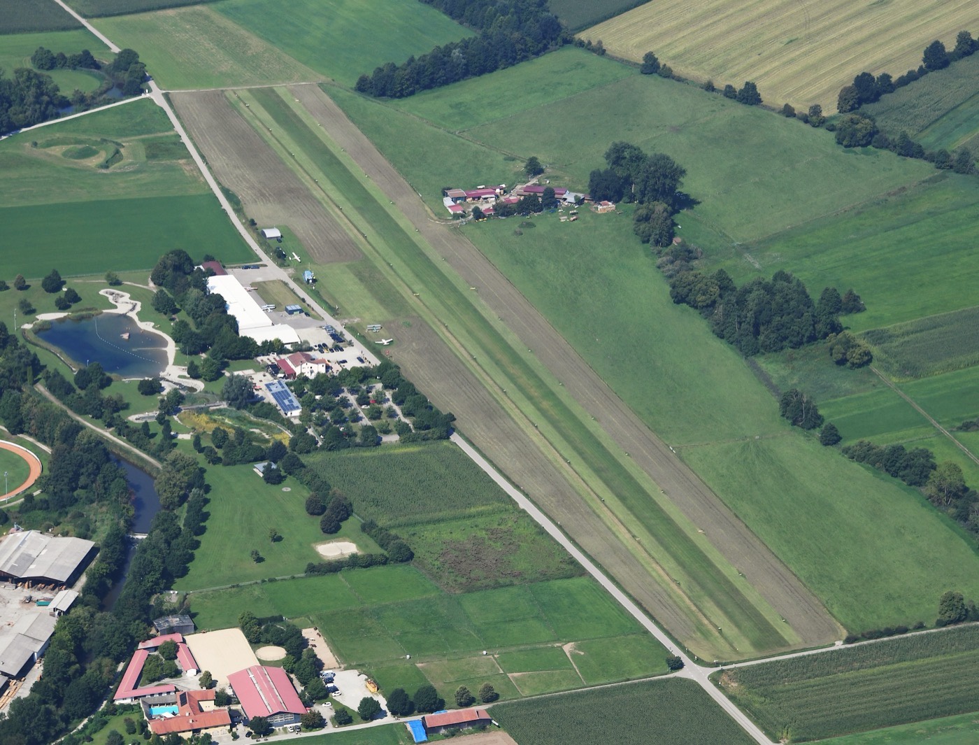 Vue aérienne verticale d'un petit aérodrome herbeux avec sa piste, ses hangars et son aire de stationnement, entouré de champs